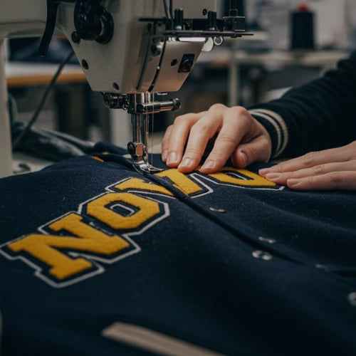 A close-up of a sewing machine stitching a navy blue varsity jacket with yellow and white 'NOLA' lettering, with a person's hands guiding the fabric.